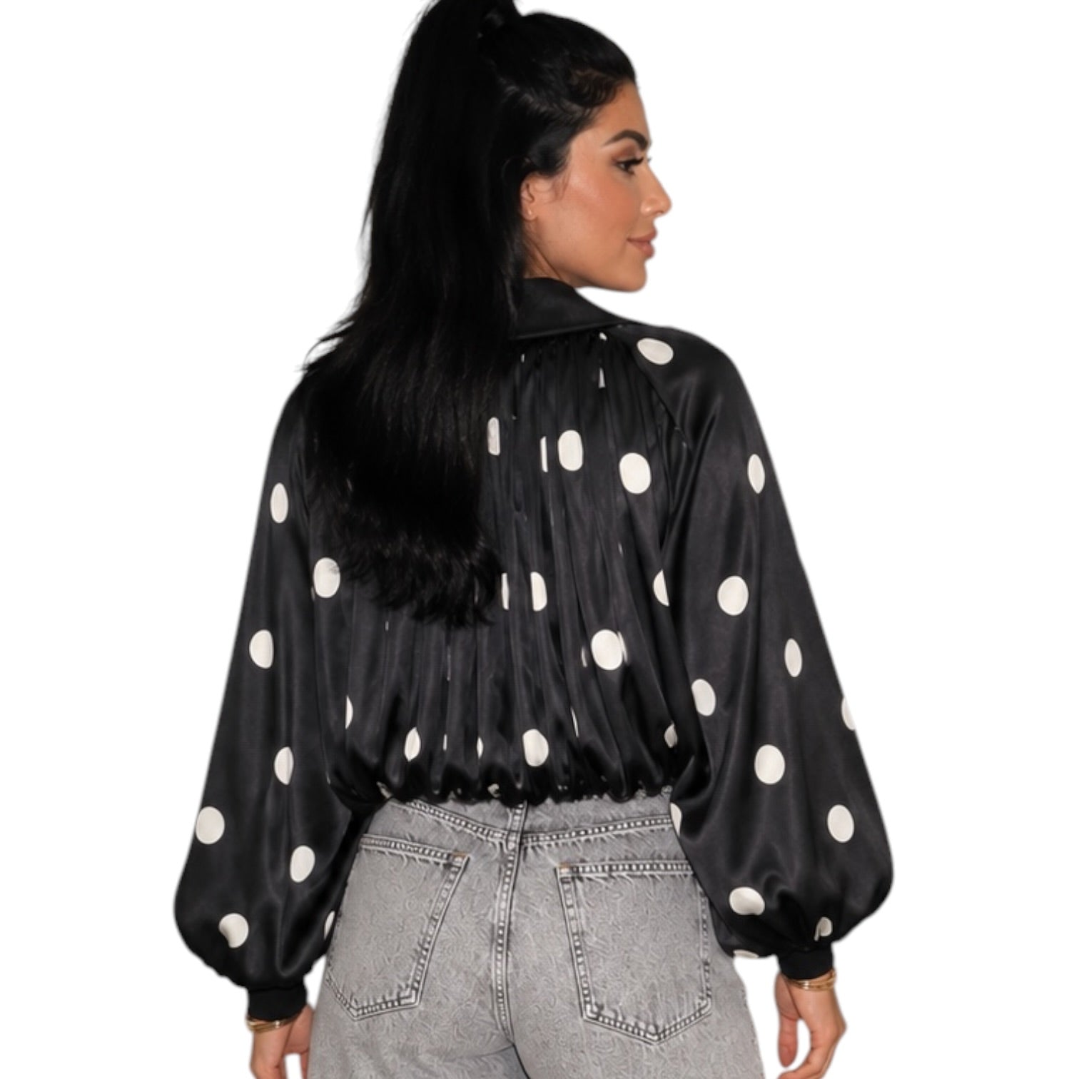 A woman shown from the back wearing a black satin-like blouse with large white polka dots and voluminous long sleeves, paired with light gray jeans, against a plain white background.