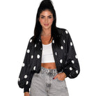 A woman modeling a black satin bomber jacket with white polka dots, worn open over a white crop top and light-wash denim jeans, against a plain white background.