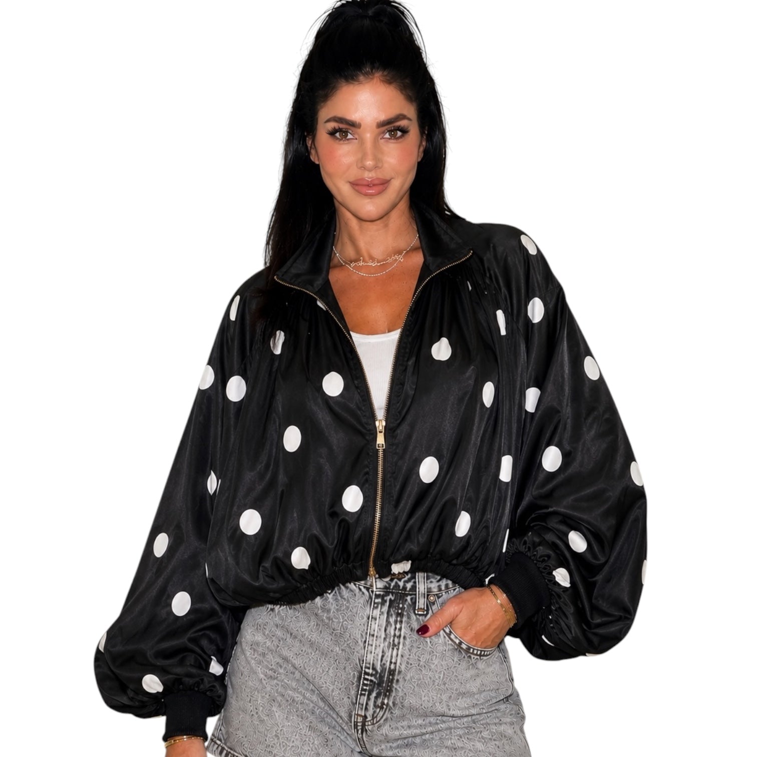 A woman wearing a black satin bomber jacket with white polka dots, styled with light-wash denim jeans and a white top, against a plain white background.