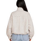 A woman viewed from the back wearing a cream-colored long-sleeve blouse with a black polka dot pattern and light-wash blue jeans, set against a plain white background.