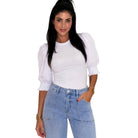 A woman modeling a white long-sleeve top with voluminous puff sleeves and ruffled cuffs, paired with light blue jeans, against a plain white background.