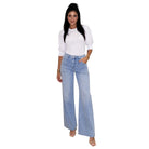A woman modeling a pair of light-wash wide-leg denim jeans with a white puff-sleeve top, standing against a plain white background.