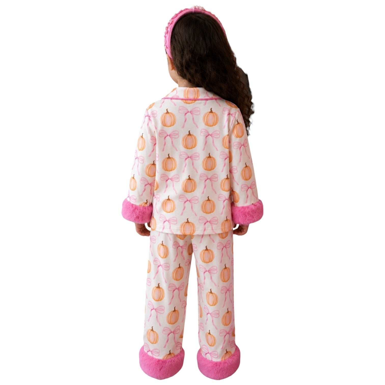 A child shown from the back wearing a matching pajama set with a pumpkin and bow print, pink trim, and a pink headband, photographed against a plain white background.