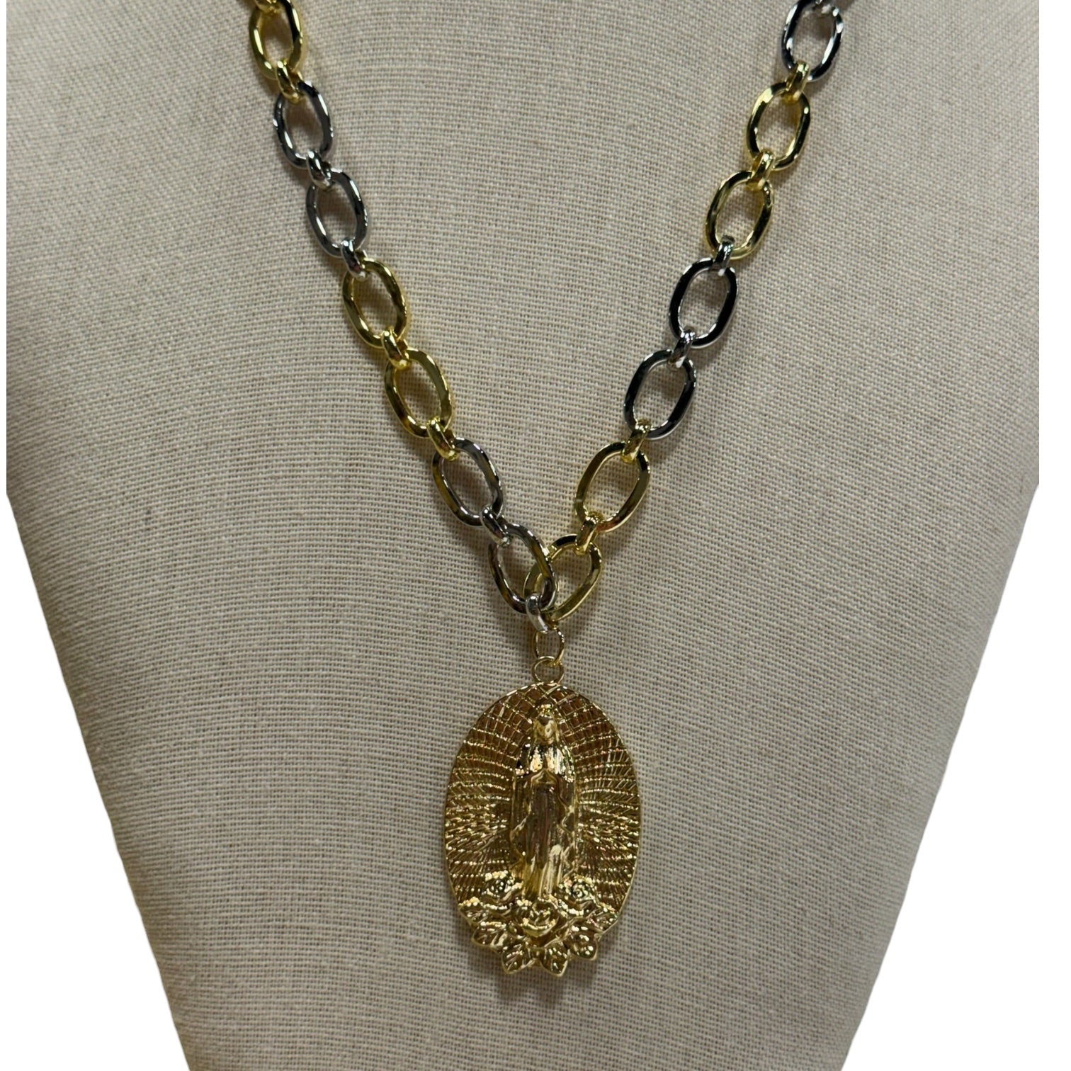 A gold and silver chain necklace with a textured gold pendant featuring an embossed religious figure, displayed on a beige fabric jewelry bust.