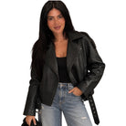 A woman wearing a black textured faux-leather moto jacket with a belted waist, styled with a black top and light-wash distressed jeans, against a plain white background.