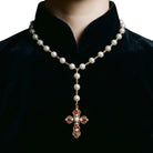 A pearl and gold cross necklace with red gemstone accents, shown worn on a person's neck over a black velvet collar.