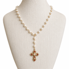 A pearl and gold necklace displayed on a cream fabric mannequin bust, featuring a long strand of white pearls with gold links and a central gold cross pendant embellished with red stones and white pearls.