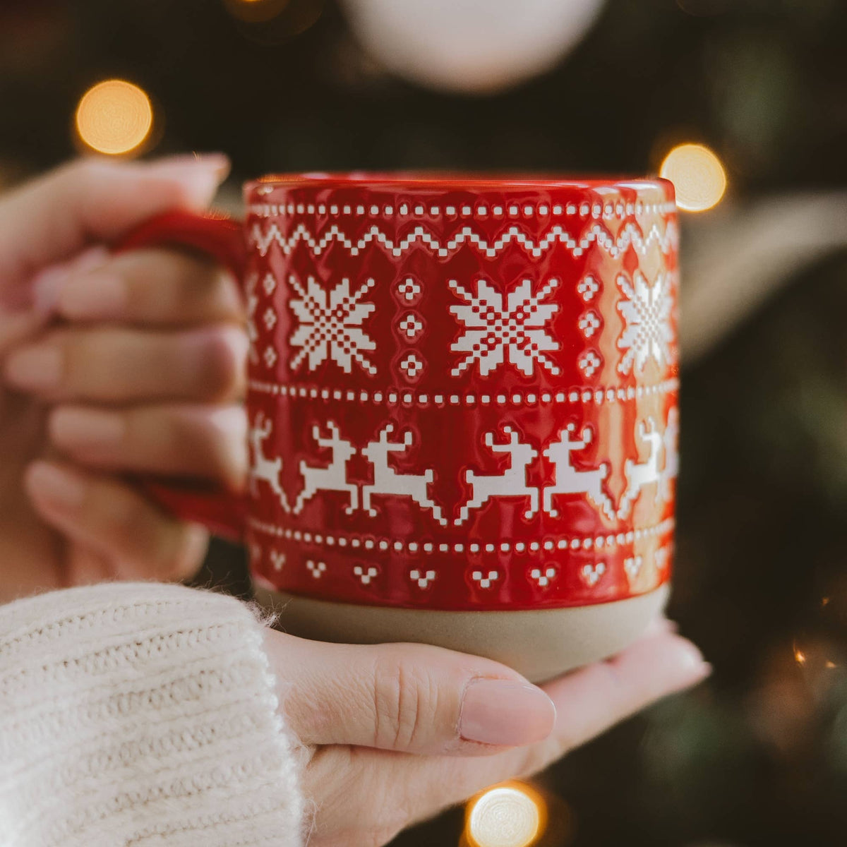A person holds a red ceramic mug with a white Nordic and Christmas pattern featuring snowflakes, reindeer, and geometric borders. The background is a warm, blurred holiday scene with soft bokeh lights.