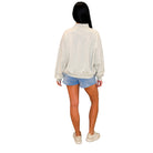 A woman shown from the back wearing a light beige or off-white long-sleeve mock-neck sweatshirt, light blue denim shorts, and off-white sneakers, set against a plain white background.