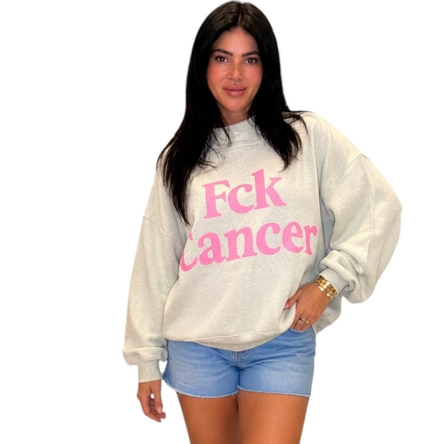 A woman modeling an oversized light gray sweatshirt with 'Fck Cancer' printed in pink across the front, paired with light blue denim shorts, against a plain white background.