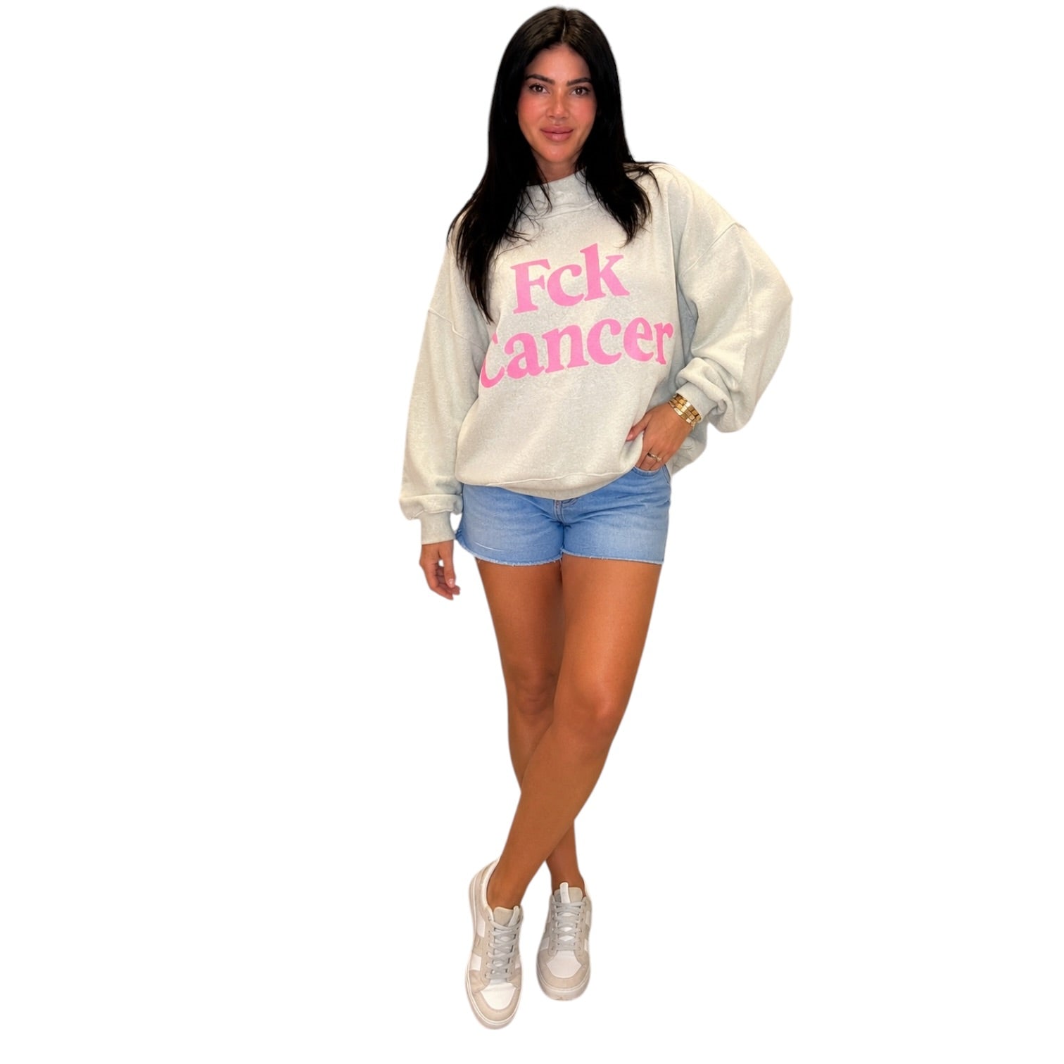 A woman modeling a cream-colored sweatshirt with 'Fck Cancer' printed in pink across the chest, paired with denim shorts and white sneakers, set against a plain white background.