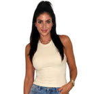 A woman with long dark hair in a high ponytail wearing a fitted cream-colored ribbed halter top and blue jeans, standing against a plain white background.