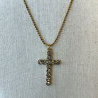A gold cross pendant necklace adorned with small clear rhinestones, displayed on a textured beige fabric background.