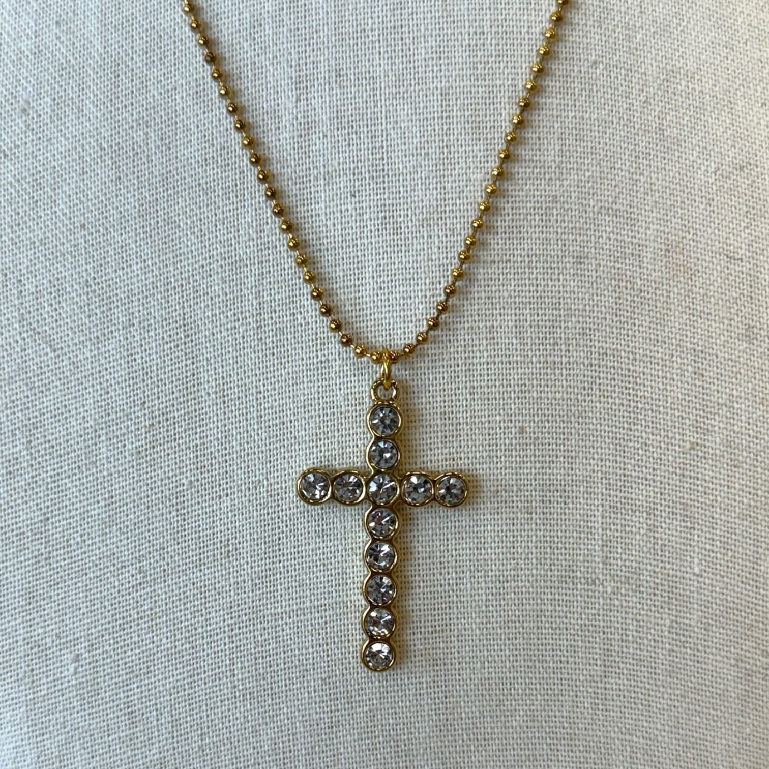 A gold cross pendant necklace adorned with small clear rhinestones, displayed on a textured beige fabric background.