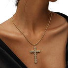 A gold cross pendant necklace adorned with clear rhinestones, worn on the neck of a model wearing a black top against a white background.