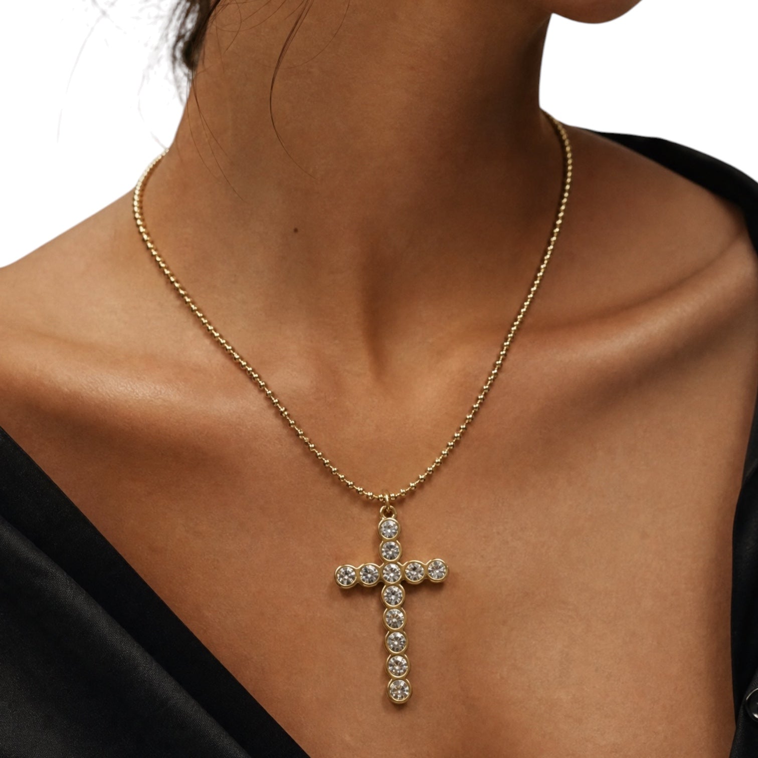A gold cross pendant necklace adorned with clear rhinestones, worn on the neck of a model wearing a black top against a white background.