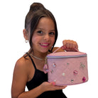 A young girl with her hair in a high bun holds a pink quilted cosmetic bag decorated with rhinestones and a dangling pink teardrop charm, against a plain white background.