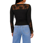 A woman shown from the back wearing a black long-sleeve top with sheer black lace panels across the shoulders and down the sleeves, paired with light blue jeans, against a plain white background.