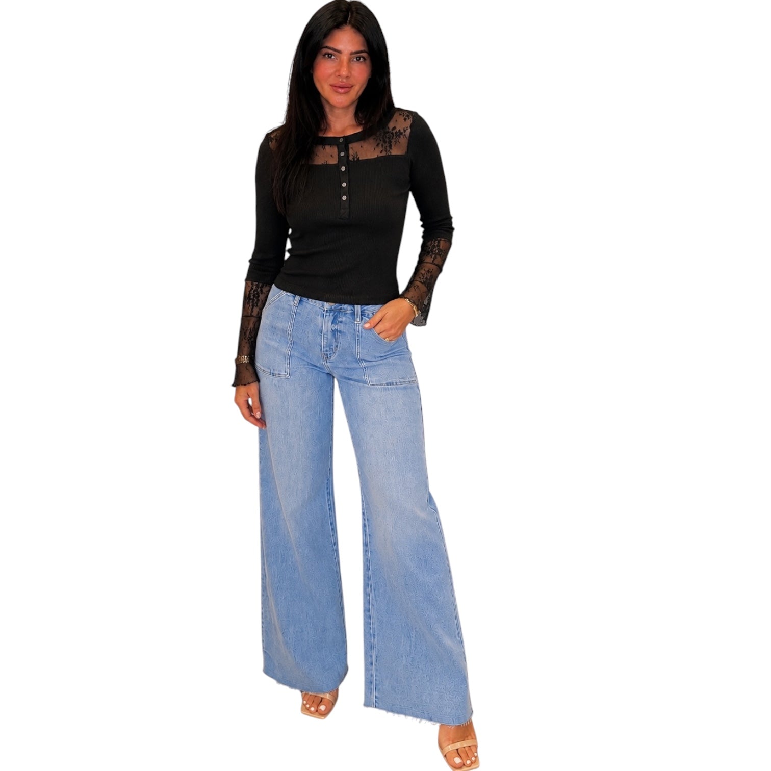 A woman modeling a black long-sleeve top with sheer lace sleeves and light-wash wide-leg denim jeans, standing against a plain white background.