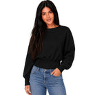 A woman with long dark hair wearing a black long-sleeve crewneck sweatshirt and blue jeans, standing against a plain white background.