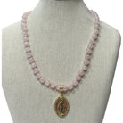 A pink stone bead necklace with a gold-toned oval religious pendant displayed on a beige fabric jewelry bust against a white background.