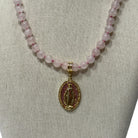 A pink stone bead necklace with a gold Miraculous Medal pendant displayed on a neutral fabric jewelry bust.