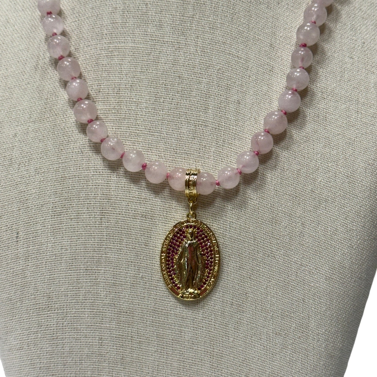 A pink stone bead necklace with a gold Miraculous Medal pendant displayed on a neutral fabric jewelry bust.