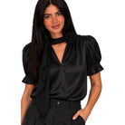 A woman modeling a black satin blouse with puff sleeves, a ruffled tie neckline, and a large fabric rose detail on the chest, paired with black trousers against a plain white background.