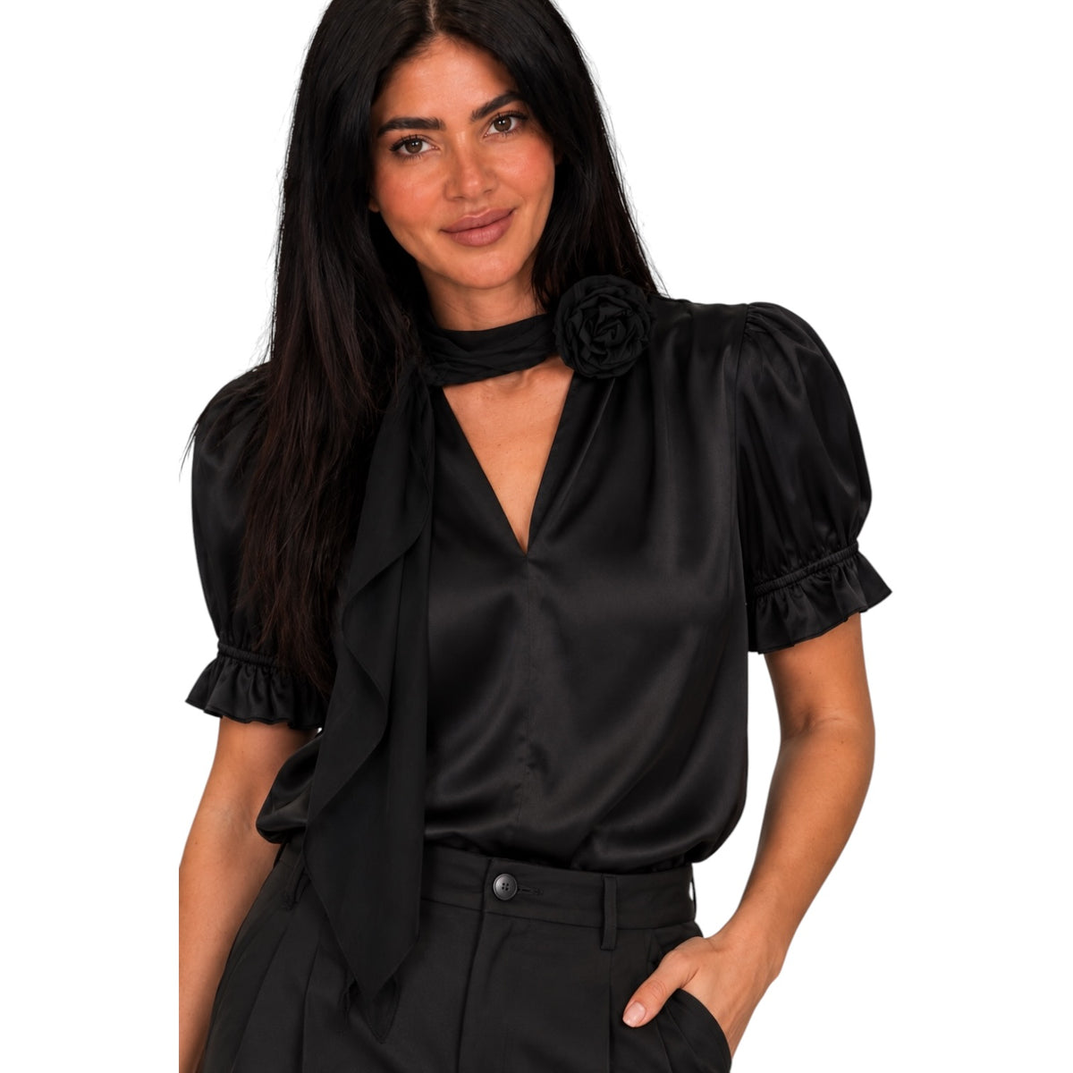 A woman modeling a black satin blouse with puff sleeves, a ruffled tie neckline, and a large fabric rose detail on the chest, paired with black trousers against a plain white background.