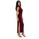 A woman modeling a sleeveless, high-neck, floor-length burgundy velvet dress with a ruched bodice and thigh-high slit, paired with black high-heeled sandals against a plain white background.