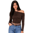 A woman modeling a dark brown off-the-shoulder long-sleeve crop top with ruched detailing, paired with blue jeans and a brown belt, against a solid white background.