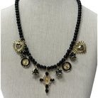 A black beaded statement necklace displayed on a neutral fabric mannequin bust. The necklace features multiple gold-toned charms, including sunburst hearts, circular medallions, small bells, and a central cross pendant embellished with gold, clear stones, and black beads.
