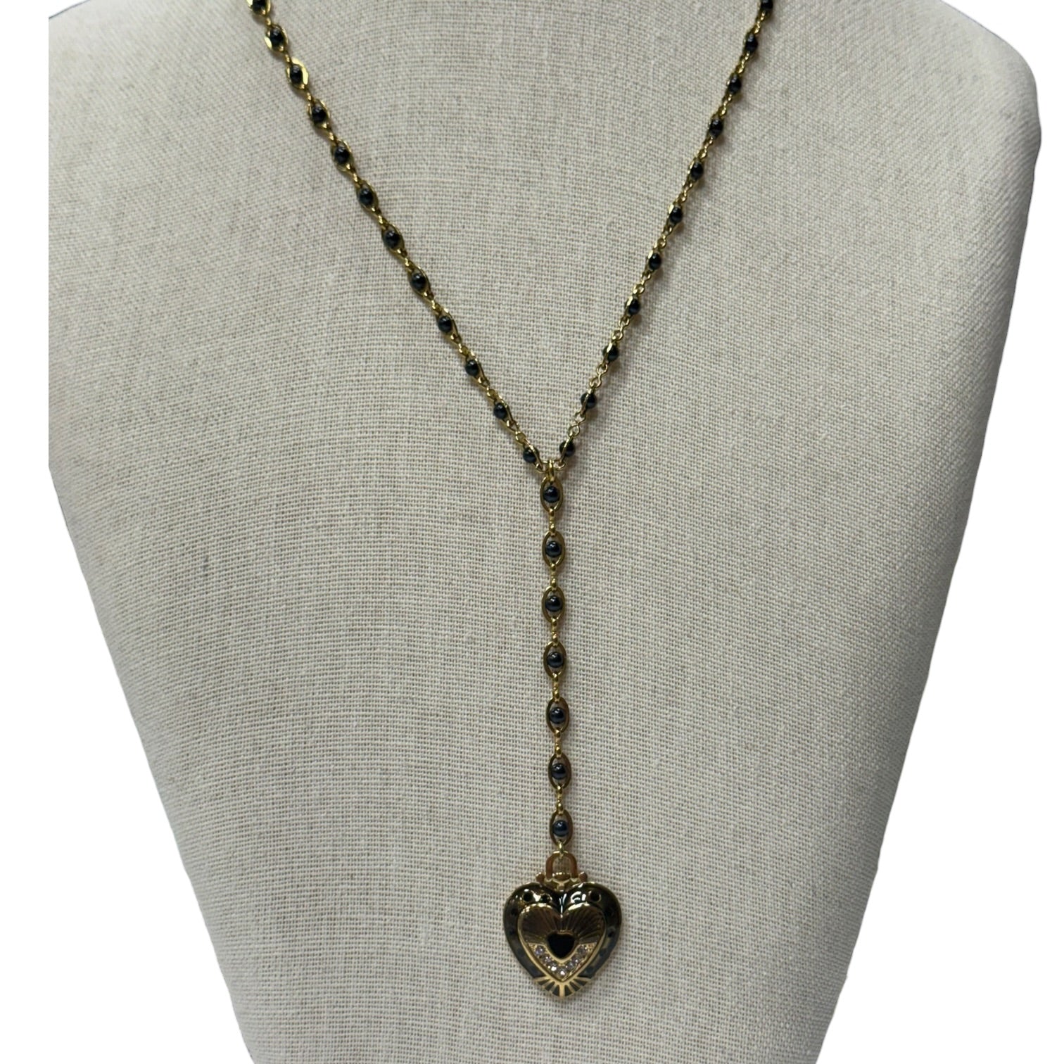 A long gold-toned beaded necklace with a heart-shaped pendant, displayed on a neutral fabric jewelry bust against a white background.
