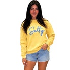 A woman modeling a yellow crewneck sweatshirt with the word 'Salty' printed in blue script across the chest, paired with light blue denim shorts, against a plain white background.