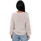 A woman viewed from the back wearing a light beige long-sleeve knit sweater with lace-patterned sleeves, paired with light blue jeans, against a plain white background.