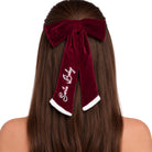 A long, dark brown velvet hair bow accessory with white faux-fur trim and the words 'Santa Baby' embroidered in white script, shown worn on a model's head from the back.
