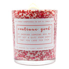 A hand-poured soy candle in a clear glass jar filled with white wax and dense pink and red glitter flakes. The jar has a white paper label with pink text that reads 'continue good' and a humorous message about showing someone who may not get a gift from Santa.