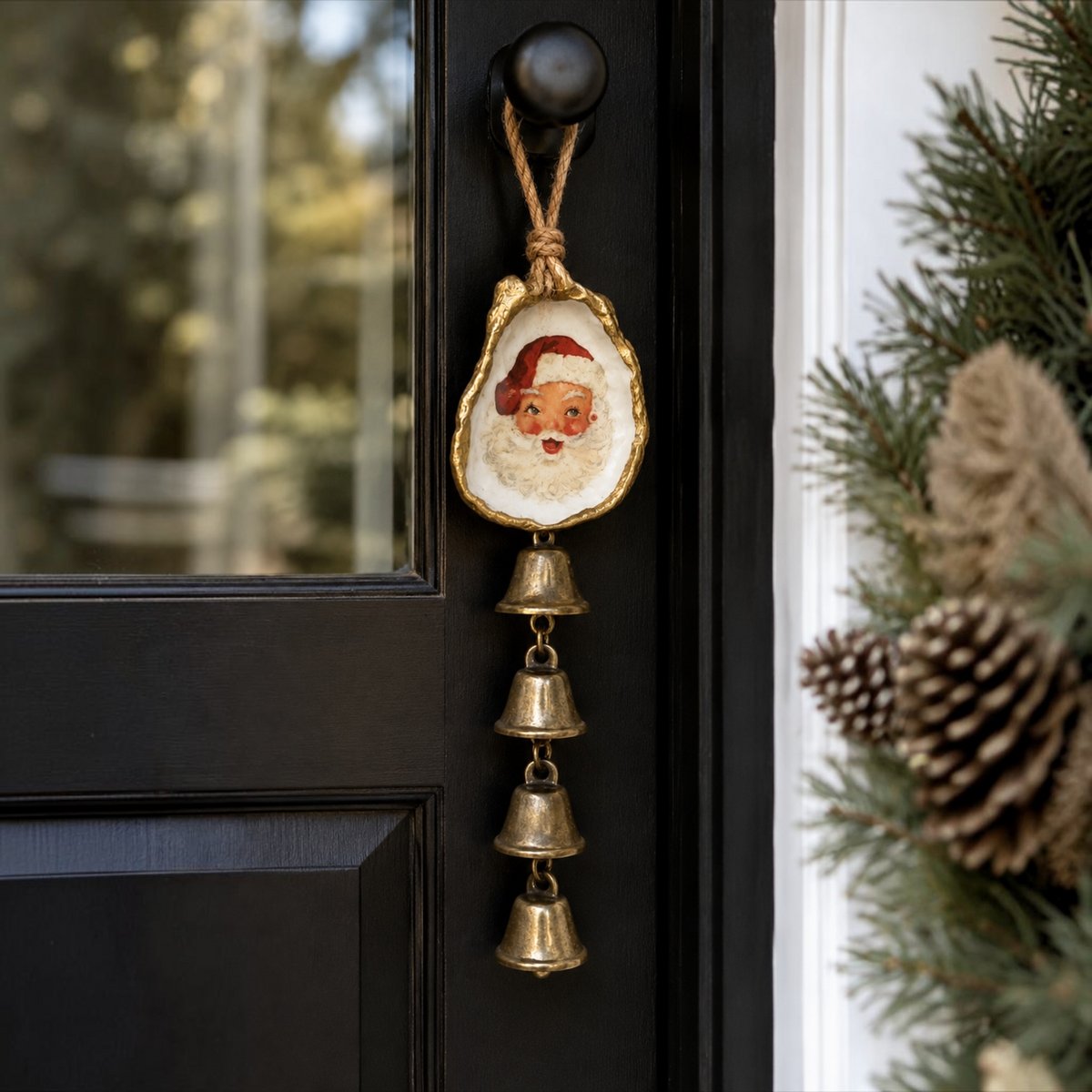 A festive Christmas door hanger ornament shaped like an oyster shell with a painted Santa face and dangling brass bells, hanging on a black front door next to holiday greenery.