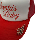 A close-up of a red and white trucker hat with 'Santa's Baby' embroidered in red script on the white front panel. The red mesh back and curved brim are also red.