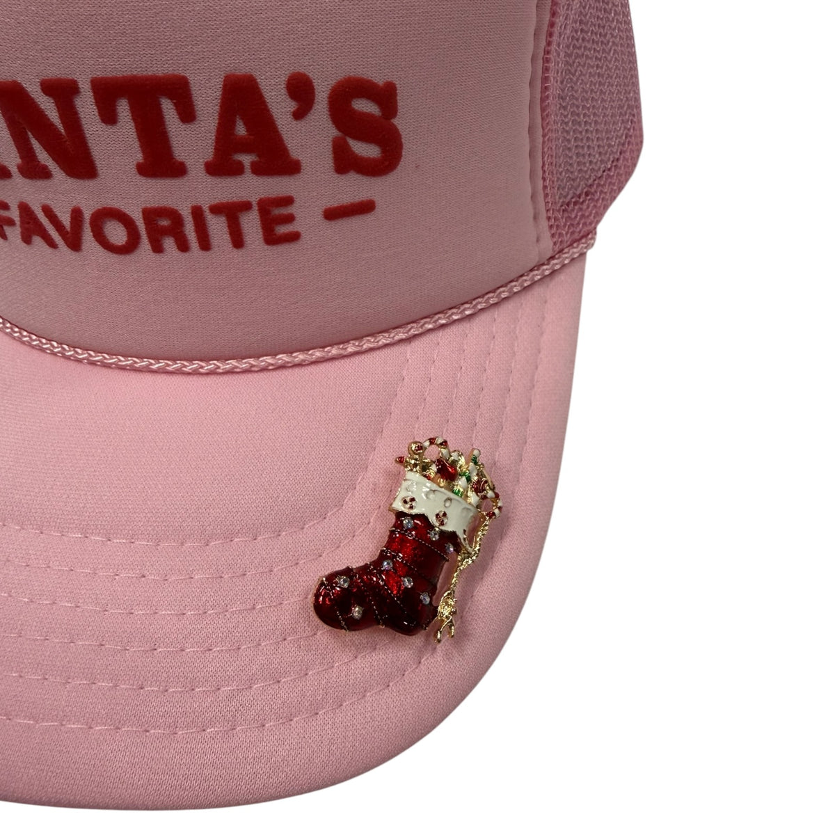A close-up of a light pink trucker hat with red embroidered text reading 'SANTA'S FAVORITE' on the front panel and a small red-and-white stocking embellishment on the brim, shown against a white background.