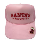 A light pink trucker-style cap with 'SANTA'S FAVORITE' embroidered in dark red on the front. The cap features a curved brim and a small red and gold stocking embellishment on the side, set against a plain white background.