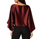 A woman shown from the back wearing a draped, wide-sleeved satin top in deep burgundy with black pants, against a plain white background.