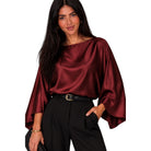 A woman modeling a satin-like boat-neck blouse in deep burgundy with wide draped sleeves, paired with black trousers and a black belt, against a plain white background.