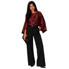 A woman modeling a satin-like burgundy top with dramatic wide sleeves and black wide-leg trousers, holding a black clutch against a plain white background.