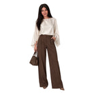 A woman modeling a cream satin blouse with wide bell sleeves and high-waisted, wide-leg brown trousers, holding a brown handbag against a plain white background.