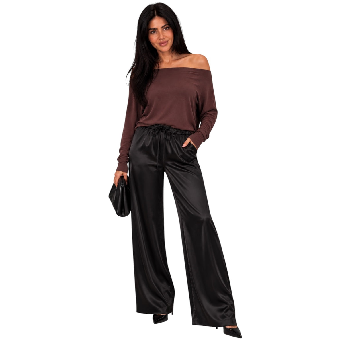 A woman modeling a two-piece outfit consisting of a brown off-the-shoulder long-sleeve top and black wide-leg satin pants, holding a small black clutch against a plain white background.
