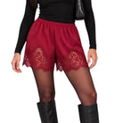 A person wearing red lace-trimmed shorts over sheer black tights, paired with a black long-sleeve top and black knee-high boots, against a plain white background.
