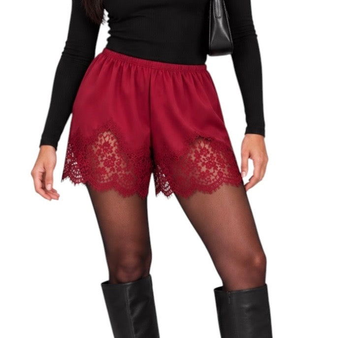 A person wearing red lace-trimmed shorts over sheer black tights, paired with a black long-sleeve top and black knee-high boots, against a plain white background.