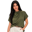 A woman modeling a draped olive green short-sleeve top with cream pants against a plain white background.