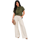 A woman modeling a short-sleeve olive green draped top and high-waisted off-white wide-leg trousers, styled with a brown belt and holding a beige clutch bag against a plain white background.